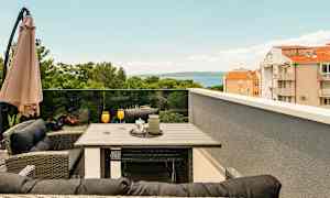 Terrasse med bord og stoler, oransje drikker i glass, parasoll og utsikt over trær, bygninger og hav. (AI-generert tekst)