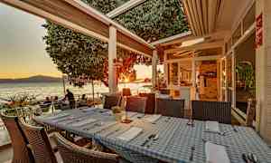 Restaurantbord med blå- og hvitrutete duker på en terrasse ved sjøen under solnedgang. (AI-generert tekst)