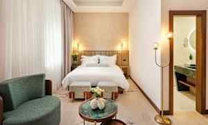 Et elegant hotellrom med en oppredd seng, lenestol og små bord. Et bad ses gjennom en åpen dør. (AI-generert tekst)