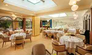 En lys og elegant restaurant med bord med hvite duker og komfortable stoler. Vinduer med planter utenfor og takvinduer gir naturlig lys. (AI-generert tekst)