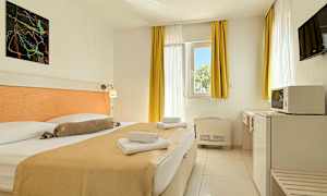 Et hotellrom med en dobbeltseng, lyse gardiner og møbler samt et vindu som slipper inn dagslys. (AI-generert tekst)