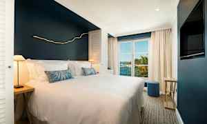 Et hotellrom med en bred seng, hvitt sengetøy og en marineblå vegg, store vinduer slipper inn naturlig lys og viser utsikt over tropisk vegetasjon og havet. (AI-generert tekst)