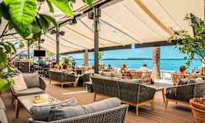 Utendørs servering med bord og sofaer, utsikt over havet i bakgrunnen. (AI-generert tekst)