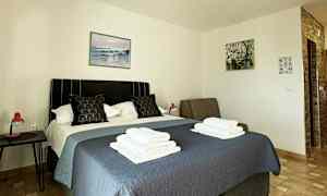 Et hotellrom med en seng dekket med et blått sengeteppe, to puter og sammenbrettede håndklær oppå. Malerier henger på veggene, og et nattbord med en rød lampe står ved siden av sengen. (AI-generert tekst)