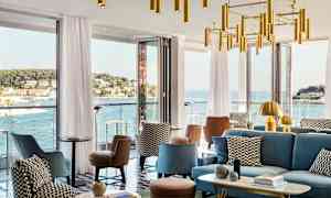 En moderne lounge med sittemøbler og dekorative lysekroner, omgitt av store vinduer som gir utsikt over en kystby og havet. (AI-generert tekst)