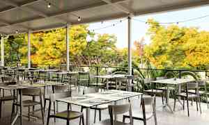 En utendørsrestaurant med bord og stoler under tak, omgitt av vegetasjon. (AI-generert tekst)