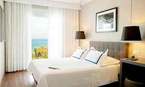 Et lyst hotellrom med en dobbeltseng, moderne innredning og havutsikt gjennom store vinduer. (AI-generert tekst)