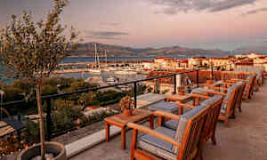 Utendørs serveringsområde med trestoler og bord, utsikt mot småbåthavn og fjell i solnedgang. (AI-generert tekst)