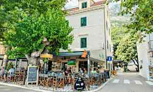 En utendørs kafé ved en bygning med hvite vegger og grønne skodder, omgitt av grøntområder og trær. (AI-generert tekst)