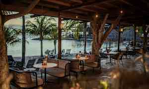 En utendørs lounge under et tretak med sofaer og bord ved havet, omgitt av palmetrær. (AI-generert tekst)