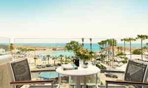 Utsikt fra en balkong med bord og stoler mot en strand med parasoller og palmer ved havet. (AI-generert tekst)