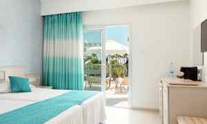 Lyst hotellrom med dobbeltseng og utsikt mot en terrasse med solsenger og parasoller. (AI-generert tekst)