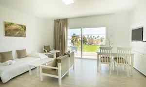 Lyst møblert rom med seng, stoler, bord og utsikt mot grønt område gjennom store vinduer. (AI-generert tekst)