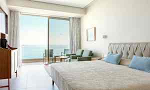 Hotellrom med seng, sittegruppe og utsikt mot havet gjennom glassdører. (AI-generert tekst)