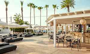 En utendørs servering med bord og stoler under et hvitt tak, omgitt av solsenger og parasoller. I bakgrunnen sees palmer og blå himmel. (AI-generert tekst)