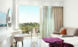 Et lyst hotellrom med en sofa, en seng med lilla teppe og en balkong med utsikt over grøntområde. (AI-generert tekst)