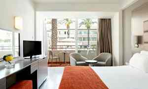 Hotellrom med seng, sitteplasser og balkong som vender ut mot en gårdsplass med palmer. (AI-generert tekst)