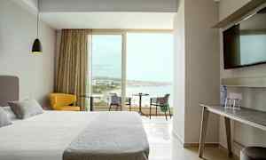 Et hotellrom med moderne innredning, en bred seng, en gul stol, et bord med utsikt over stranden gjennom store vinduer. (AI-generert tekst)