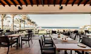 Restaurant med uteservering, palmer og hav i bakgrunnen. (AI-generert tekst)