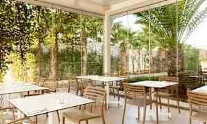 Bilde av en uteservering med bord og stoler omgitt av glassvegger og grøntområder. (AI-generert tekst)