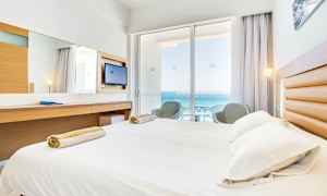 Hotellrom med seng, skrivebord og balkong med utsikt over havet. (AI-generert tekst)