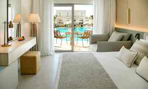 Et hotellrom med seng, sofa og utgang til en balkong med utsikt mot bassengområdet. (AI-generert tekst)