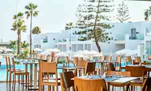 Restaurant med trestoler og dekte bord, utsikt mot hvit arkitektur, basseng og palmer. (AI-generert tekst)