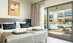 Et hotellrom med en dobbeltseng, lysegrå sengegavler og blå puter. Et bilde med iskremmotiv henger på veggen. Gjennom glassdøren vises et utendørsbasseng med solsenger og parasoller. (AI-generert tekst)