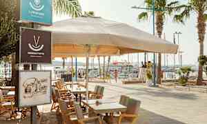 Uteområde med bord og stoler under en stor parasoll ved en havn. (AI-generert tekst)