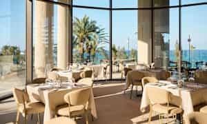 En lys og elegant restaurant med panoramavinduer som viser utsikt over palmer og havet. (AI-generert tekst)