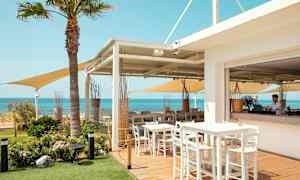 En utendørsrestaurant med bord og stoler, utsikt over havet og klar blå himmel. (AI-generert tekst)