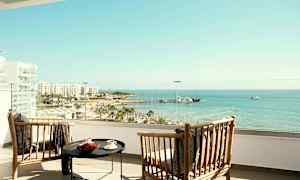 To trestoler og et bord på en hotellbalkong med utsikt over strand og hav. (AI-generert tekst)