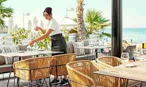 Uteservering ved sjøen med palmer i bakgrunnen. (AI-generert tekst)