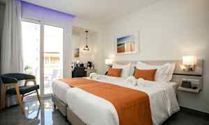 Et lyst hotellrom med to enkeltsenger oppredd med hvitt sengetøy og oransje dekorelementer. Det er en balkong med glassdører, en stol og et skrivebord med speil. (AI-generert tekst)