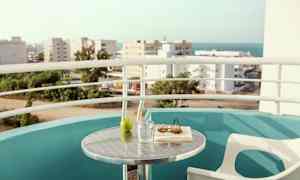 En balkong med utsikt over by og hav. Et bord med et glass, et grønt eple, en bok og solbriller. (AI-generert tekst)