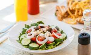 En tallerken med salat som inneholder tomat, agurk, løk, fetaost og oliven. I bakgrunnen sees pommes frites og smørbrød. (AI-generert tekst)