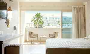 Et hotellrom med en seng, skrivebord og stol ved siden av en balkong med utsikt over bymiljø. (AI-generert tekst)