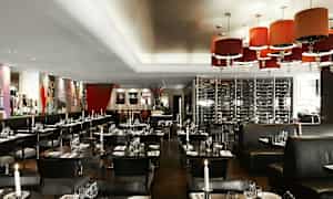 En elegant innredet restaurant med tente lys, mørke møbler og et hengende vinkjeller som bakgrunn. (AI-generert tekst)