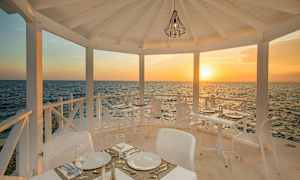 En utendørsrestaurant med hvite møbler, plassert ved sjøen, med solnedgang i bakgrunnen. (AI-generert tekst)