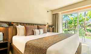 Et hotellrom med en stor seng utstyrt med flere puter, nattbord og en glassdør som fører til en uteplass og grønne omgivelser. (AI-generert tekst)