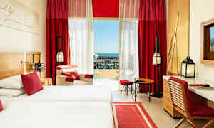 Et hotellrom med to senger, en sitteplass, et skrivebord og røde gardiner ved balkongen. (AI-generert tekst)