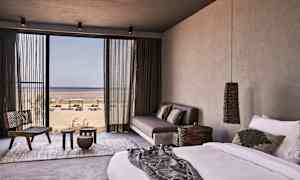 Et hotellrom med seng, sofa og utsikt mot stranden gjennom en stor glassdør. (AI-generert tekst)