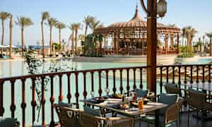 Et dekket bord på en veranda med utsikt over et basseng omgitt av palmer og en paviljong. (AI-generert tekst)