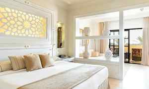 Et lyst og elegant soverom med dekorerte vaser på en hylle, en stor seng med puter og utgang til en balkong. (AI-generert tekst)