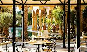 Utendørs serveringsområde med bord og stoler under et tak, omgitt av vegetasjon og palmer. (AI-generert tekst)