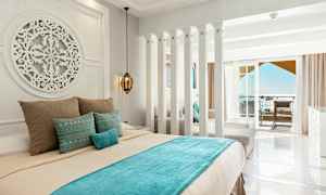 Et hotellrom med lyse vegger, en dekorert seng med blå og beige sengetøy, samt en balkong med havutsikt. (AI-generert tekst)