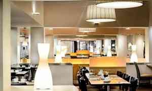 En elegant spisesal med moderne interiør, der bord er dekket med servise og bestikk. Belysningen består av tak- og bordlamper. (AI-generert tekst)