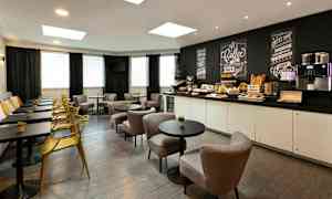 Et caféområde med bord, stoler og en benk langs veggen. Et serveringsbord med frokosttilbehør og en kaffemaskin er synlig i bakgrunnen. (AI-generert tekst)