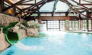 Et innendørsbasseng med en vannsklie nær en klippe og et kunstig fossefall. Bassengområdet er omgitt av store vinduer og trekonstruksjoner. (AI-generert tekst)