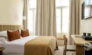 Hotellrom med en dobbeltseng, puter og et sengeteppe i brune toner samt et skrivebord med en kaffemaskin. (AI-generert tekst)